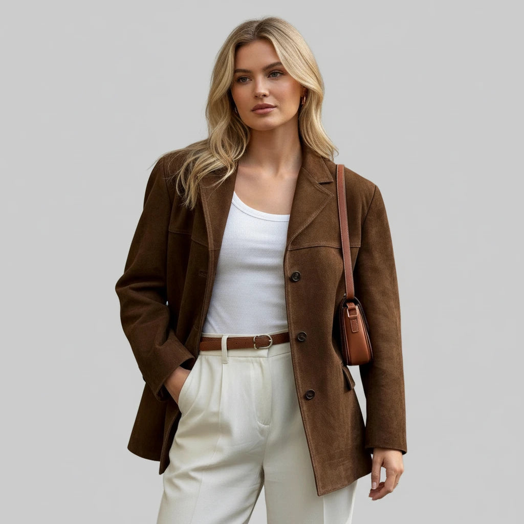 Woman wearing a brown coat and white pants with a brown bag on a gray background