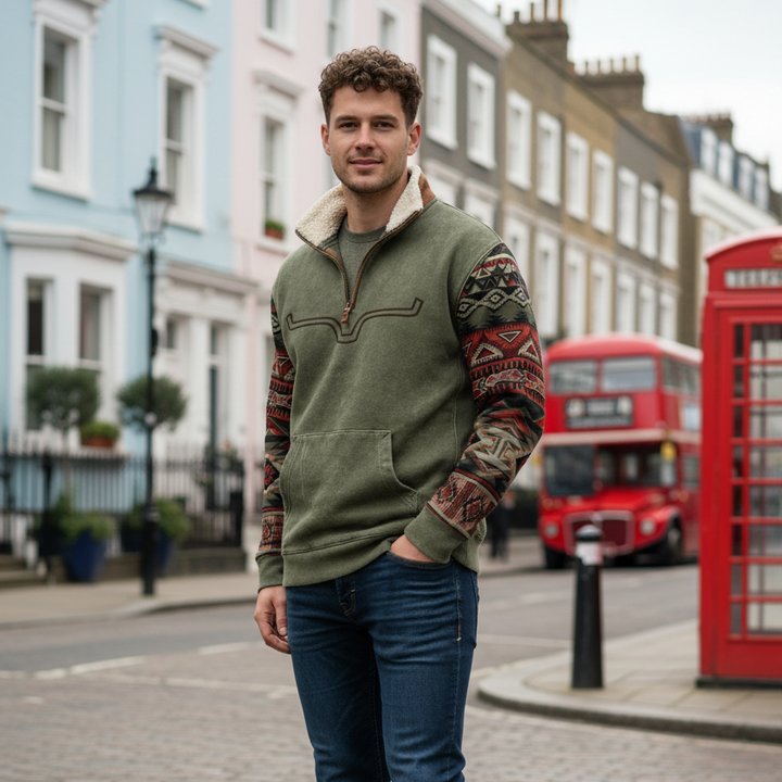 Man wearing a green fleece jacket with patterned sleeves standing on a street with a red double-decker bus in the background.